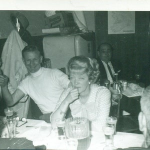 Mulher de 1965 sentada à mesa de festa, marido muito bêbado ao lado da foto dos anos 60, preto e branco