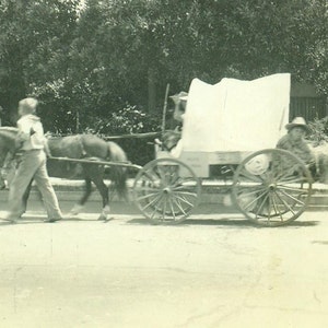 May include: A black and white photo of a horse-drawn wagon with a white canvas top. The wagon is being pulled by two horses and is driven by a person sitting on a seat in the front. There are people standing around the wagon, and a person on horseback in the background.