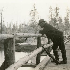 Cacciatore dell'Alaska alla ricerca di salmoni nel ruscello in piedi su un ponte di tronchi di legno con il fucile appoggiato accanto a lui Foto antica Fotografia in bianco e nero