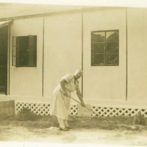 May include: A black and white photo of a woman standing in front of a house. She is holding a rake and is wearing a long-sleeved shirt and pants. The house is a simple, one-story structure with a porch and a few windows.