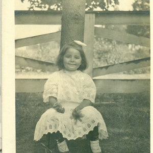 Petite fille assise à l'extérieur tenant un cookie et une robe à œillets blancs des années 1910 Antique RPPC Real Photo Carte postale