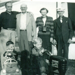 May include: Black and white vintage photograph of six people, including two young children. The children are playing with toy vehicles. The adults are standing together, smiling. The image has a nostalgic feel, likely taken in the mid-20th century.