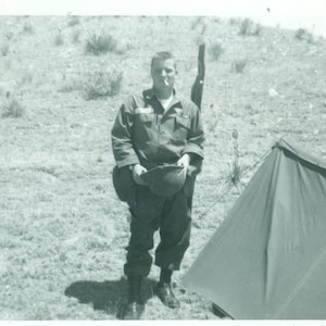 May include: A man in military uniform stands in a field holding a helmet and a rifle. He is standing near a tent.
