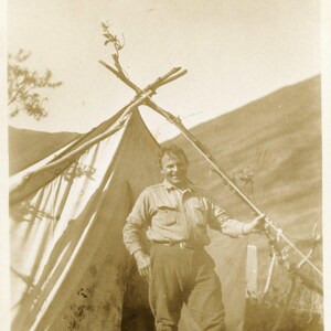 Fotografia antica di un uomo cacciatore dell'Alaska degli anni '20 che campeggia in piedi fuori dalla tenda