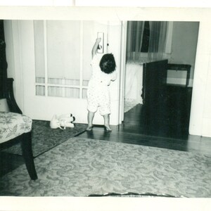 May include: A black and white photo of a young child in a white and black patterned shirt standing in a doorway. The child is reaching for a door handle. The photo is taken from a low angle.