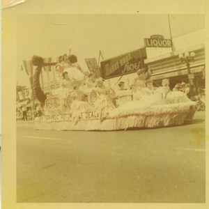 May include: A vintage photograph of a parade float decorated with flowers and featuring a group of women in white dresses. The float is adorned with the text "Marks Bros. Shoes" and "San Fernando Auto & Le Deal."