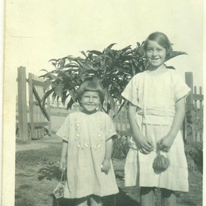 Op de afbeelding: Twee jonge meisjes staan voor een houten hek en een bladplant. Het meisje links draagt een witte jurk met een bloemenpatroon en zwarte schoenen. Het meisje rechts draagt een witte jurk met een gerimpelde kraag en zwarte schoenen.