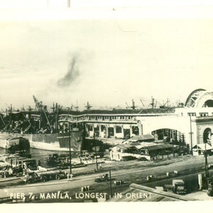 Może przedstawiać: Zdjecie w stylu vintage czarno-białe przedstawiające Pier 7 w Manili na Filipinach, z napisem "Longest in Orient". Obraz przedstawia tętniący życiem port z statkami, budynkami i różnymi pojazdami. Dym unosi się w tle.