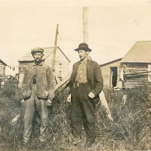 Antico Akutan Alaska Uomini in piedi fuori Foto Estate Capanne di tronchi Abiti da lavoro Fotografia d'epoca