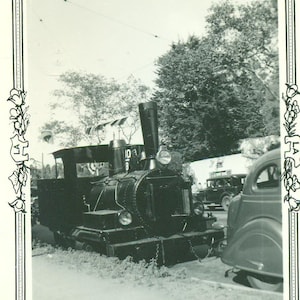 May include: A black and white photo of a small steam locomotive pulling a car. The locomotive has a number "108" on the side. The car is a vintage sedan.