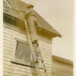 Uomo d'epoca dell'Alaska in piedi su una scala che dipinge il tetto di una casa che guarda verso la telecamera RPPC cartolina fotografica antica