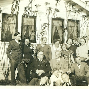 May include: A vintage black and white photograph of a large family posing in front of a house. The group includes men in suits and women in dresses, with several children. The house has multiple windows and is decorated with plants.