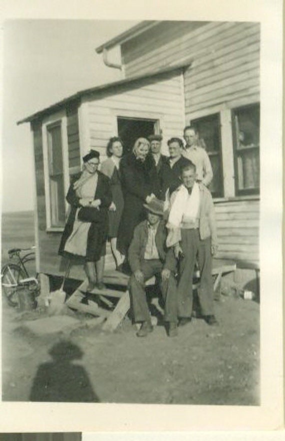 Gathered on weathered wooden steps, this multi-generational family creates a heartwarming tableau against their humble clapboard home, their faces glowing with genuine joy and connection. The simple architecture and casual 1940s attire tell a story of resilience and togetherness during simpler times ✨ Notice how the photographer's shadow in the corner adds an intimate, documentary feel to this precious family moment.