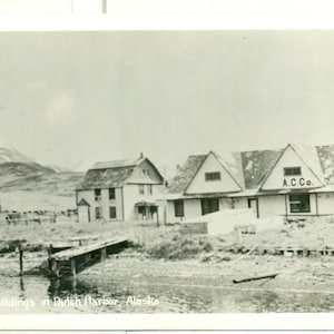 Pode incluir: Fotografia a preto e branco de edifícios em Dutch Harbor, Alasca. A imagem mostra vários edifícios com telhados inclinados, alguns com o texto "A.C. Co.". Uma estrutura de madeira estende-se para a água em primeiro plano.