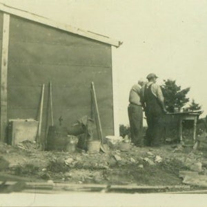 May include: A black and white photo of three people standing in front of a wooden building. The people are wearing casual clothing and are looking at something off camera. There are tools and other items in the background.