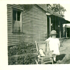 Poupée Baby Sitting en osier Chaise berçante Fille debout à l’extérieur de la ferme 1910s Antique RPPC Real Photo Carte postale Photographie