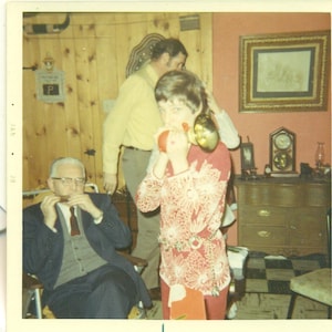 Puede incluir: Una fotografía vintage captura una reunión familiar. Una persona con una blusa floral roja toca una bocina de fiesta. Una persona mayor con un traje toca la armónica. Otra persona está de pie en el fondo. La habitación tiene paneles de madera y una imagen enmarcada.