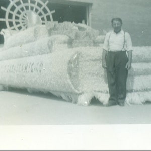 May include: A black and white photograph of a man standing in front of a large, elaborate float decorated with white flowers. The float has the words "TELEPHONE CO." written on it.