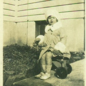 May include: A young child wearing a white coat and hat sits on a toy tricycle. The tricycle is red and has a black seat and wheels.