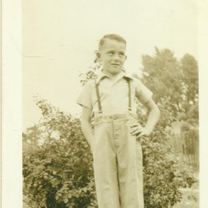 Könnte beinhalten: Ein kleiner Junge in einem weißen Hemd mit Hosenträgern und heller Hose steht vor einem Busch. Er schaut nach rechts aus dem Bild.