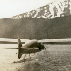 Puede incluir: Imagen en blanco y negro de un hidroavión en el agua, con una montaña de fondo. La aeronave tiene una sola hélice y flotadores. La escena evoca la aviación temprana.