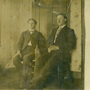 Brüder junge Männer sitzen im Haus Flasche zwischen ihnen Tapete HolzBöden RPPC Postkarte antike Foto