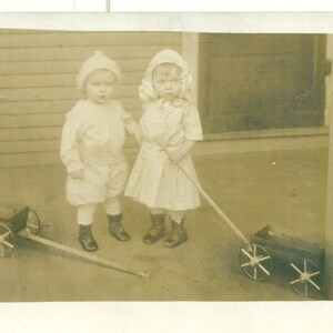May include: Two children in white dresses and hats stand in front of a wooden porch. They are holding a wooden wagon with black wheels. The wagon is pulled by a stick.