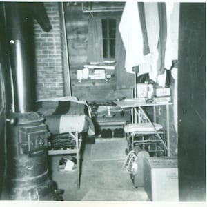 May include: A black and white photo of a room with a wood-burning stove, a bed, and a table with a chair. The room is sparsely furnished and has a rustic feel.