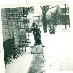 May include: A vintage black and white photograph shows a child shovelling snow. The child is wearing a plaid jacket and a hat. The date "JAN 56" is visible on the left side of the photo.
