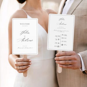 May include: Two white wedding fan programs with wooden handles. The fans have a light gray border and feature elegant calligraphy with the names "Helen & Andrew." One fan says "Welcome" and the other has a wedding timeline.