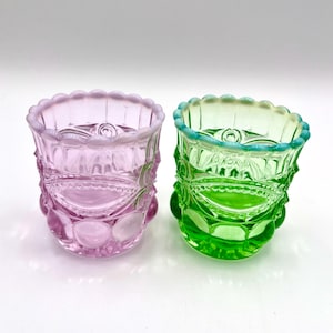 May include: Two glass votive holders with a scalloped rim and a textured pattern of raised circles and oval shapes. One holder is light pink and the other is bright green. Both pieces feature a translucent finish and a repeating geometric design.