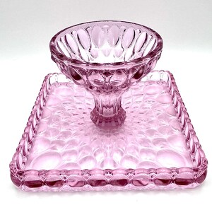 Op de afbeelding: Een roze glazen serveerset met een voetbalkom en een vierkant dienblad. De kom en het dienblad hebben een gestructureerd patroon van ovale vormen. De set is gemaakt van transparant glas en is geschikt voor het serveren van desserts of hapjes.