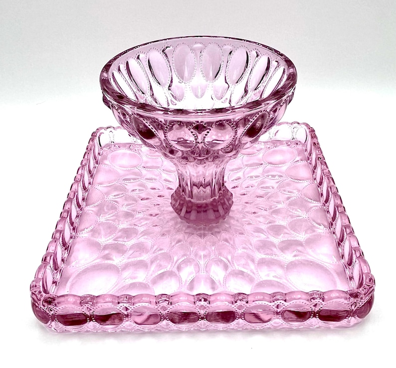 Op de afbeelding: Een roze glazen serveerset met een voetbalkom en een vierkant dienblad. De kom en het dienblad hebben een gestructureerd patroon van ovale vormen. De set is gemaakt van transparant glas en is geschikt voor het serveren van desserts of hapjes.