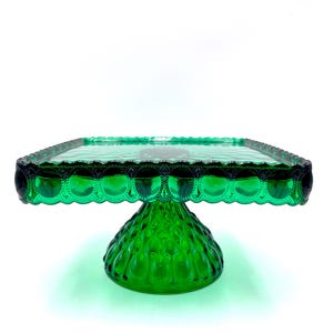 Op de afbeelding: Een heldergroene glazen taartstandaard met een vierkante bovenkant en een decoratieve geschulpte rand. De basis heeft een getextureerde, afgeronde vorm. De standaard is ontworpen voor het presenteren van taarten en desserts.