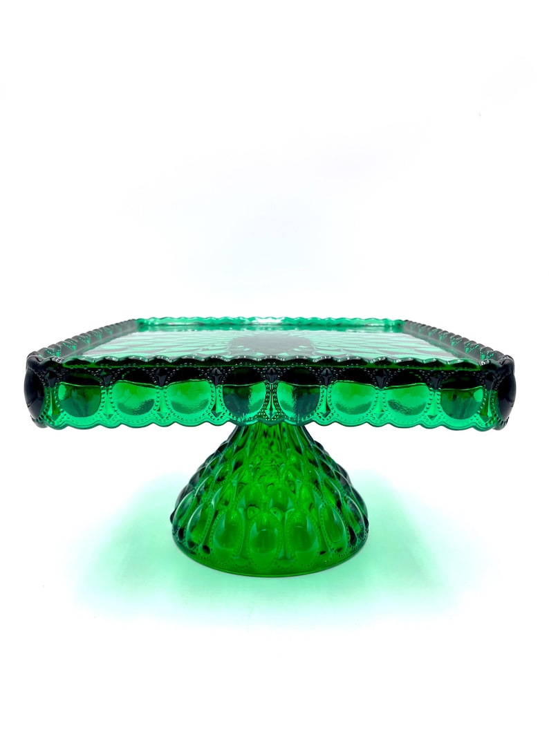 Op de afbeelding: Een heldergroene glazen taartstandaard met een vierkante bovenkant en een decoratieve geschulpte rand. De basis heeft een getextureerde, afgeronde vorm. De standaard is ontworpen voor het presenteren van taarten en desserts.