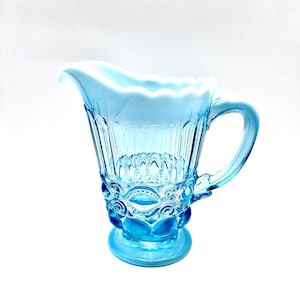 May include: A light blue glass jug with a white rim and intricate embossed designs. The jug has a rounded base and a handle.