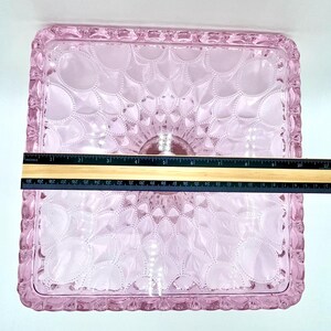Op de afbeelding: Een vierkant, roze glazen dienblad met een decoratief bloemmotief. De lade heeft een verhoogde, getextureerde rand. De lade meet ongeveer 25 cm in lengte en breedte.