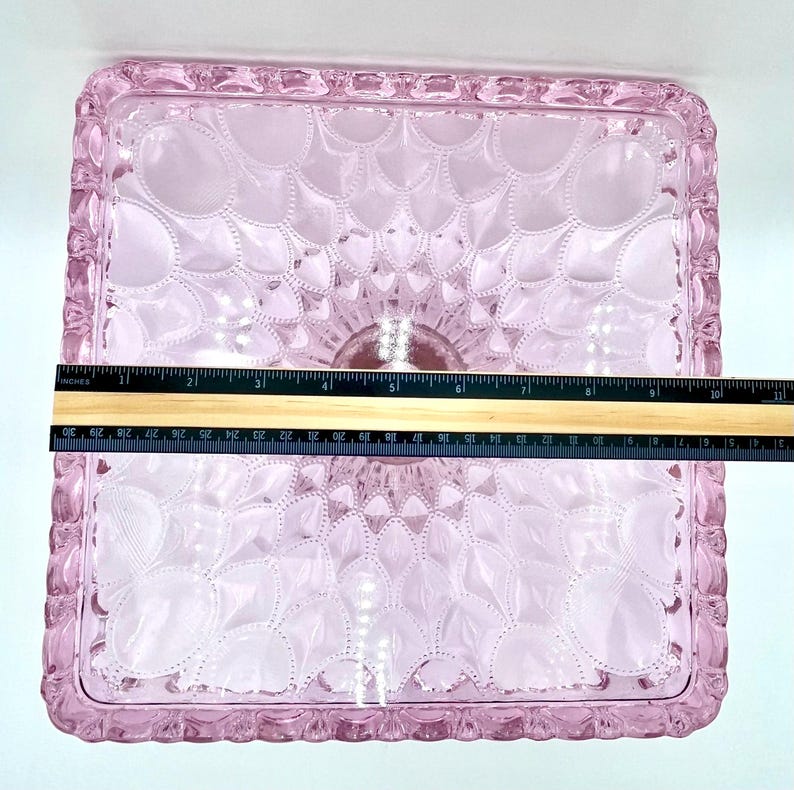 Op de afbeelding: Een vierkant, roze glazen dienblad met een decoratief bloemmotief. De lade heeft een verhoogde, getextureerde rand. De lade meet ongeveer 25 cm in lengte en breedte.