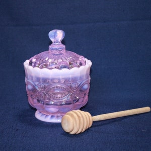 May include: A pink glass honey pot with a white rim and a wooden honey dipper. The pot has a decorative design and a lid.