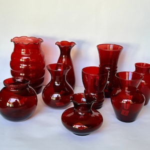 May include: A selection of nine ruby red glass vases in diverse shapes and sizes. Some vases feature ruffled rims, while others are smooth. The vases are arranged on a white surface, reflecting light and creating highlights.