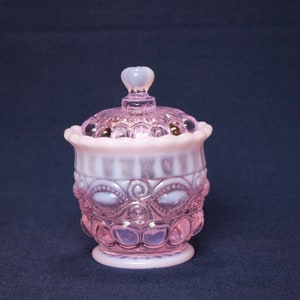 May include: A pink, lidded glass jar with detailed embossed patterns. The jar has a round body with a decorative design and a matching lid with a small, rounded handle. The jar is made of opalescent glass.
