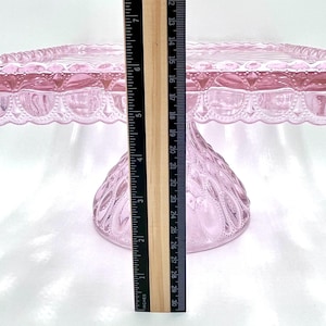 Op de afbeelding: Een roze glazen taartstandaard met een vierkante bovenkant en een decoratieve geschulpte rand. De standaard is ongeveer 20 cm hoog. De basis heeft een textuur met een diamantachtig patroon. Een liniaal is naast de standaard geplaatst.