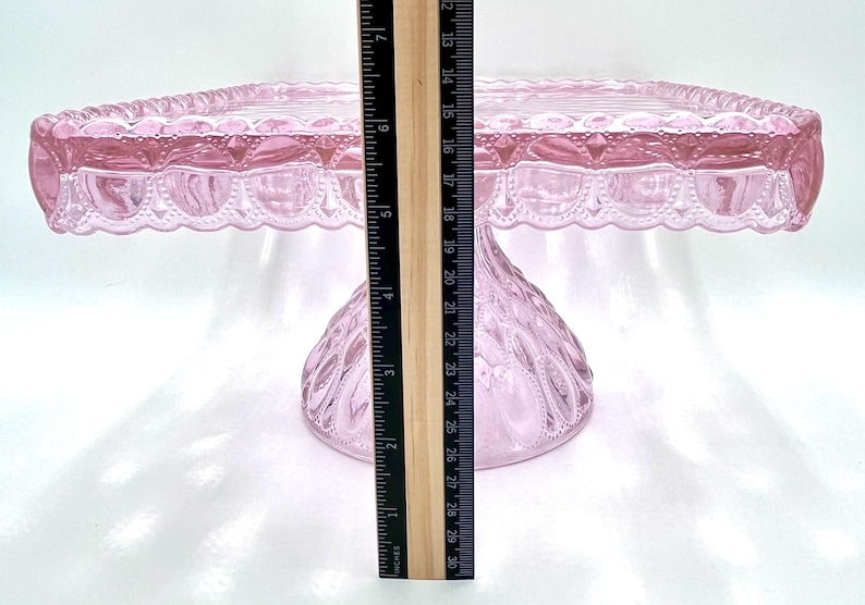 Op de afbeelding: Een roze glazen taartstandaard met een vierkante bovenkant en een decoratieve geschulpte rand. De standaard is ongeveer 20 cm hoog. De basis heeft een textuur met een diamantachtig patroon. Een liniaal is naast de standaard geplaatst.