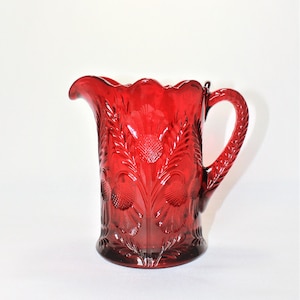 Peut inclure: Un pichet en verre rouge avec un motif de chardon. Le pichet a une large ouverture et une poignée.