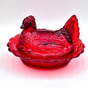 May include: A ruby red hen-shaped covered dish. The dish features a detailed hen design with textured feathers and a scalloped base. The lid is shaped like the hen's body and head. A decorative kitchenware item.