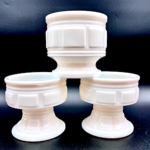 Peut inclure: Trois coupes sur pied en verre opalin blanc disposées en pile, avec une coupe placée au-dessus de deux autres. Chaque coupe présente un motif répétitif de blocs rectangulaires autour du bol et une base évasée avec un design géométrique.