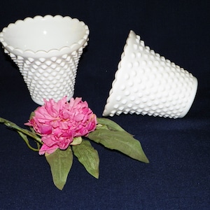 May include: Two white milk glass bud vases with a textured, raised dot pattern. The vases are empty and are sitting on a dark blue surface. A pink flower is in the center of the image.