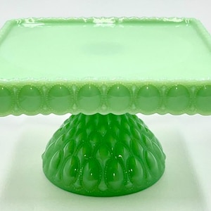 Op de afbeelding: Een groene glazen taartstandaard met een vierkante bovenkant en een decoratieve geschulpte rand. De basis heeft een afgeronde vorm met een textuurpatroon. De standaard is lichtgroen.