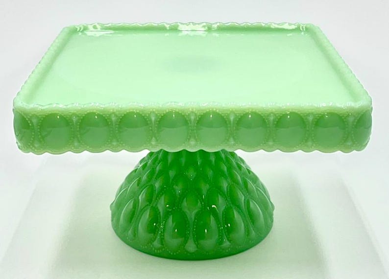 Op de afbeelding: Een groene glazen taartstandaard met een vierkante bovenkant en een decoratieve geschulpte rand. De basis heeft een afgeronde vorm met een textuurpatroon. De standaard is lichtgroen.
