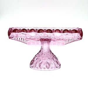 Op de afbeelding: Een roze glazen taartstandaard met een vierkante bovenkant en een decoratieve, geschulpte rand. De standaard heeft een gedetailleerde, getextureerde basis en steel. De standaard is gemaakt van glas en is ideaal voor het presenteren van taarten en andere desserts.